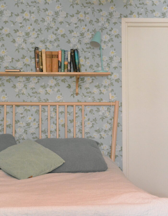 Makuuhuone, sinisävyinen kuviollinen tapetti, valkoinen ovi oikealla, puinen sänky, sängyn päällä puinen hylly. Talo Järvenpäässä Uudellamaalla.