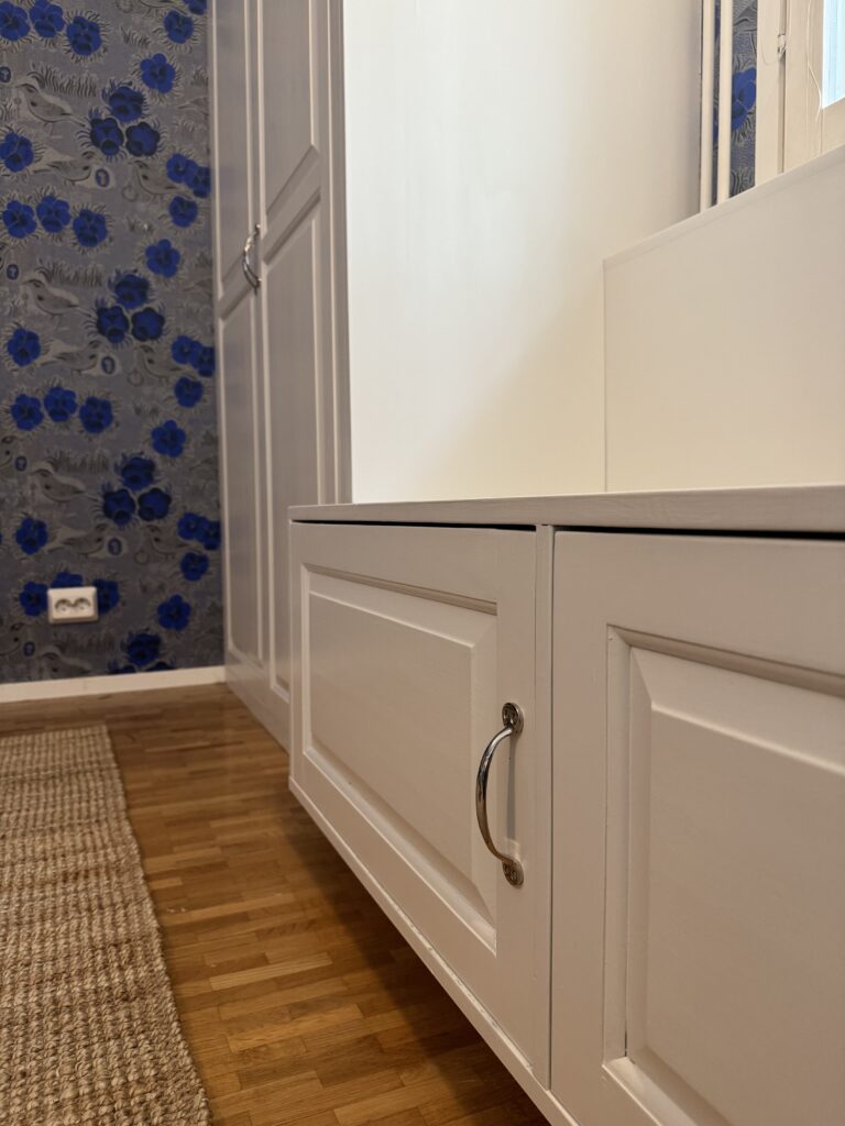 Valkoinen kiintokaluste ikkunan edessä talossa Järvenpäässä. Takana näkyy värikäs tapetti. Puuseppä Uudeltamaalta.